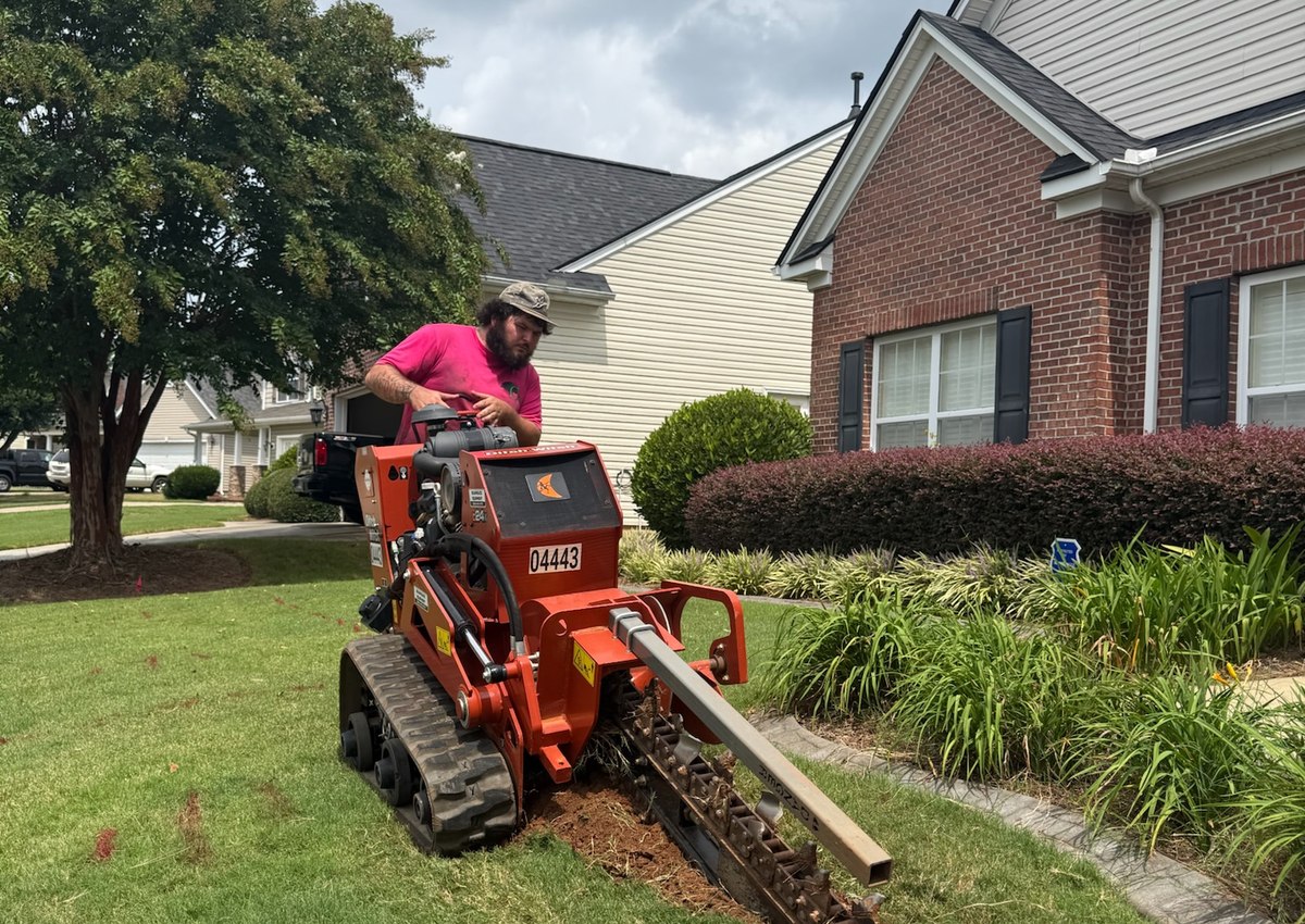 A crew member running a walk-behind trencher across a front lawn to lay irrigation line.