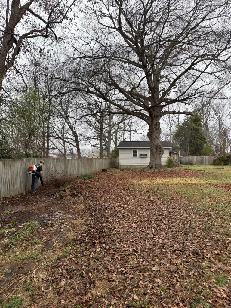 A bare back yard before work, covered in leaves and packed dirt with a crew member clearing along the fence.