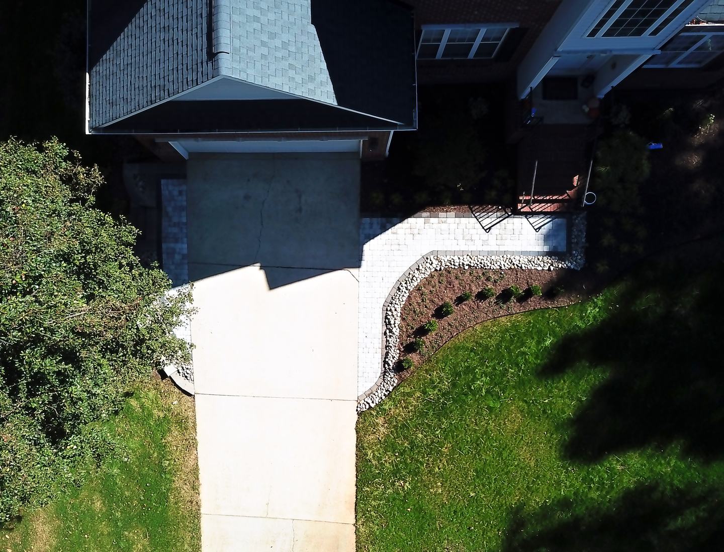 From above: a finished paver walkway running from the driveway to the front steps, edged in river rock, with a new planted bed and green lawn.