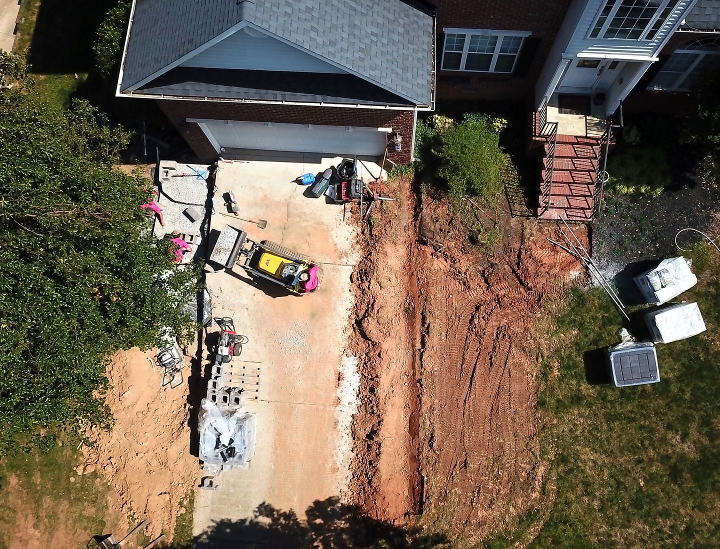 The same driveway from above before the work: a dug-out trench of red clay beside the drive, with equipment and pallets of block staged.