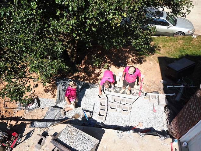 Crew members in pink shirts setting retaining wall block on a gravel base beside a driveway, seen from above.