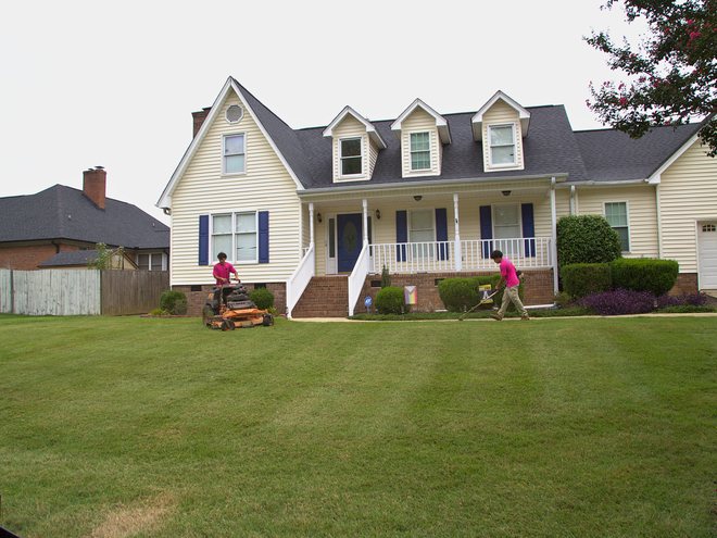 A crew mowing a wide front lawn in front of a two story home.
