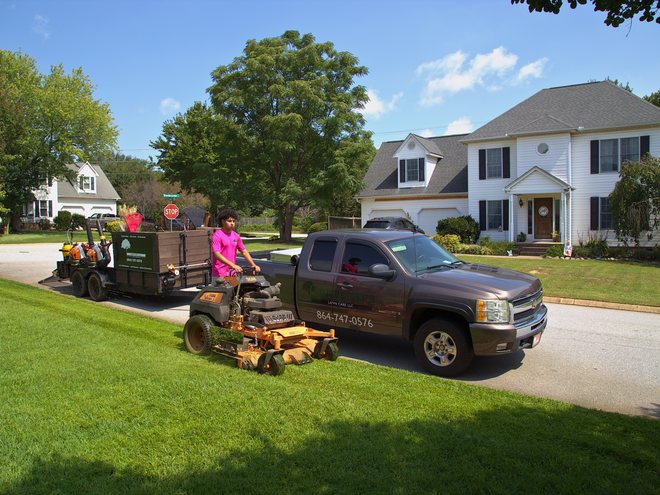 A crew member on a stand-on mower beside the company truck and trailer on a sunny day.