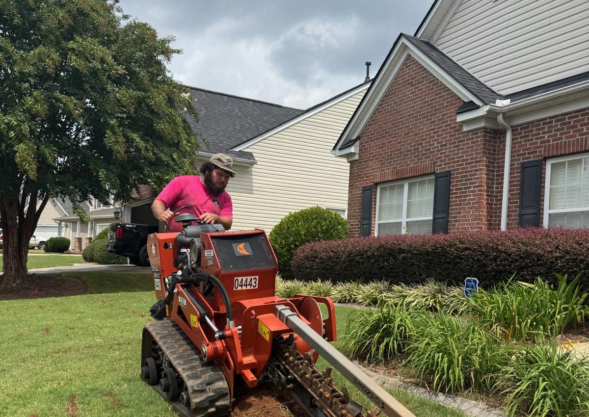 A trencher cutting a line for new irrigation pipe beside a driveway.