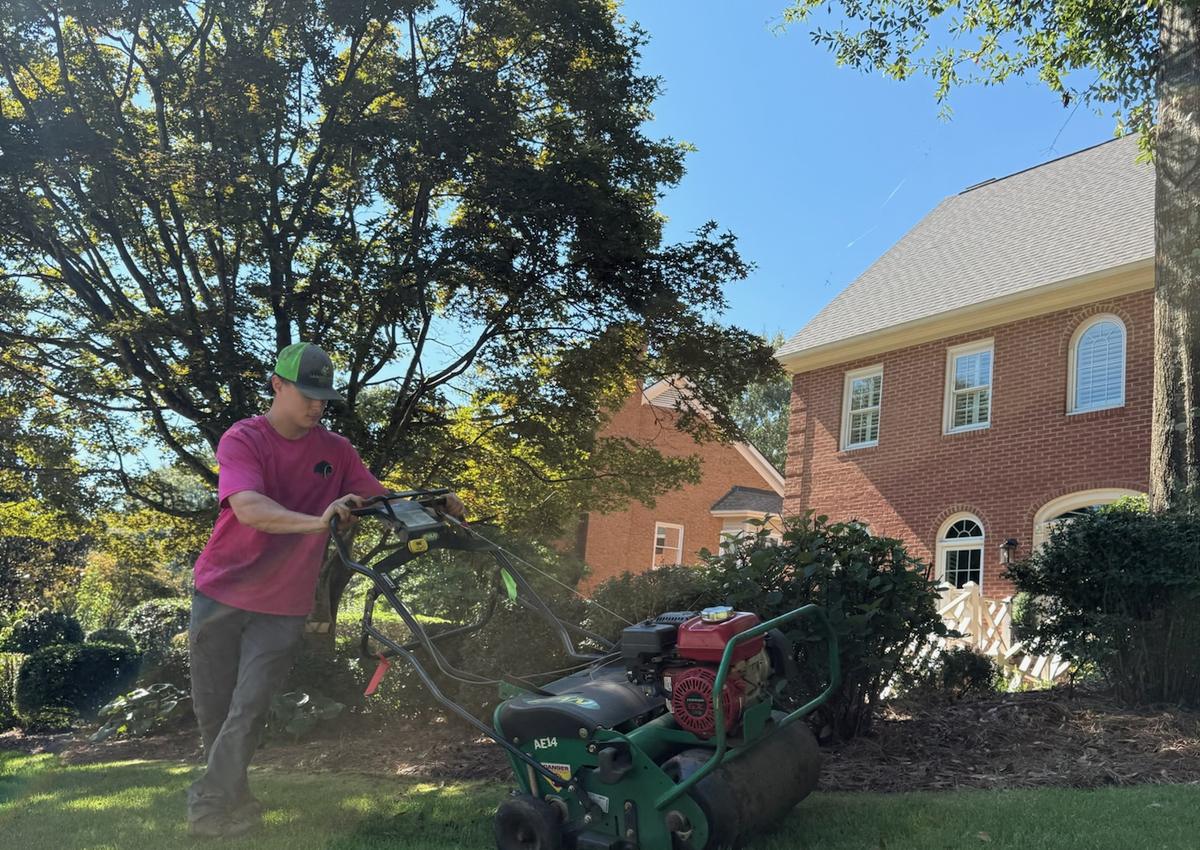 A crew member running an aeration machine across a residential lawn.