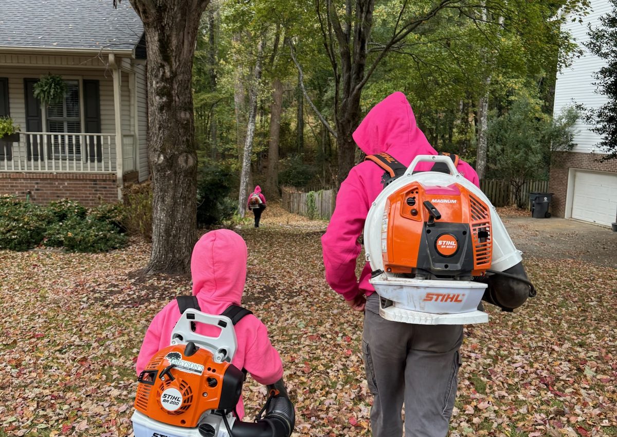 Two crew members in fuchsia hoodies clearing a leaf covered yard with backpack blowers.